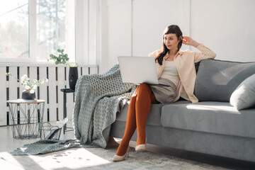 A young woman works on a laptop, communicates online in a modern bright office on a sofa on a sunny day.