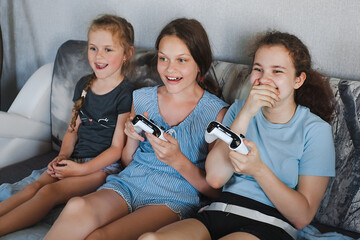 Pretty little girls are playing game console and laughing while sitting on sofa at home