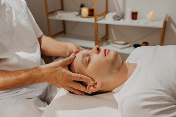 Fototapeta na wymiar Young man relaxing during professional head massage in SPA salon. Manual migraine treatment. Concept of wellness, body and health care.