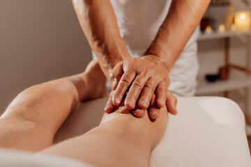 Professional legs massage in SPA salon on the background of candles. Handsome masseur therapist in white uniform making manual therapy for athlete muscles. Concept of wellness, body and health care.