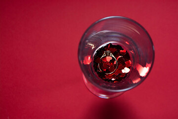Wedding ring and confetti of their red hearts in a champagne glass on a red background. Top of view