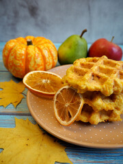 Belgian gluten-free pumpkin waffles lie on a brown plate with a dry slice of lemon and stars of anise, pumpkin, apples against a background of yellow maple leaves on a blue wooden table. vegan