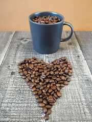 a dark gray mug filled with roasted whole coffee beans stands on a wooden background with coffee beans laid out in the shape of a heart. side view. energy