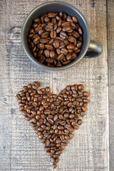a dark gray mug filled with roasted whole coffee beans stands on a wooden background with coffee beans laid out in the shape of a heart. energy