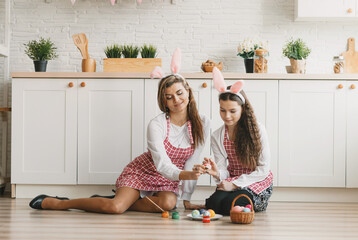 two beautiful girls are sitting on the kitchen floor and painting Easter eggs with a brush.