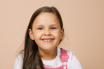 Portrait of child with bright shining eyes and excited happy smile with upper teeth looking at camera wearing bright, pink jumpsuit and white t-shirt on beige background.