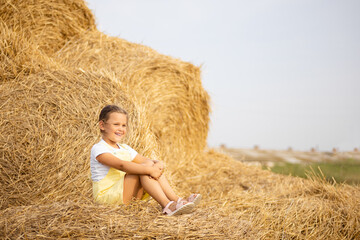 Naklejka na ściany i meble Shining Caucasian little girl calmly sitting on haystack looking at someone with teeth smile and happy face. Time away from city in country field with tons of hayricks.