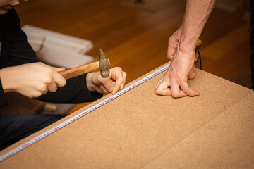 Close up of male hands measuring length of plywood with tape measure and female hands hammering nail into cheapboard. Assembling piece of furniture at home. Copy space. Fixing tools.
