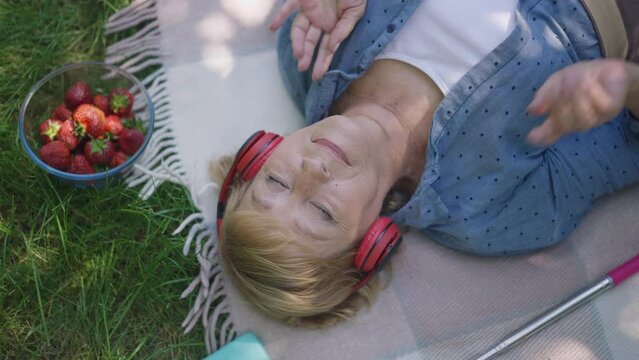 Relaxed Senior Woman Listening To Music In Headphones Lying On Blanket In Sunny Summer Spring Park. Top View Happy Carefree Caucasian Retiree Enjoying Hobby Outdoors Dancing In Slow Motion