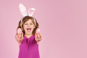 Laughing little girl with bunny ears headband holds easter eggs in her hands and covering her eyes with them. Copy space