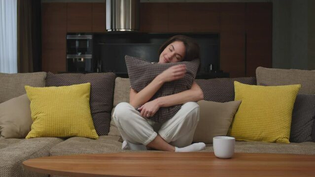Calm young woman smiling hugs pillow sitting on cozy sofa in living room at home, resting, enjoying weekend