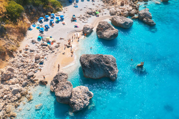 Aerial view of blue sea, sandy beach with umbrellas, stones and rocks in water at sunny day in summer. Lefkada island, Greece. Tropical landscape with sea coast, swimming people, blue water. Top view