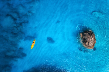 Aerial view of yellow kayak in blue sea at sunset in summer. Man on floating canoe in clear azure water, rocks, stones. Lefkada island, Greece. Tropical landscape. Sup board. Active travel. Top view