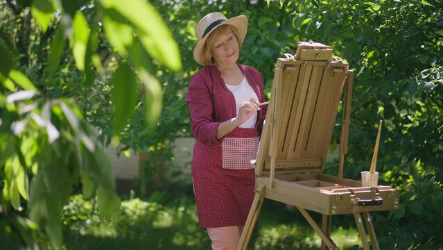 Portrait of talented senior Caucasian woman painting in slow motion on easel standing in sunny garden park. Concentrated confident retiree enjoying hobby outdoors in sunshine. Fine art and aging