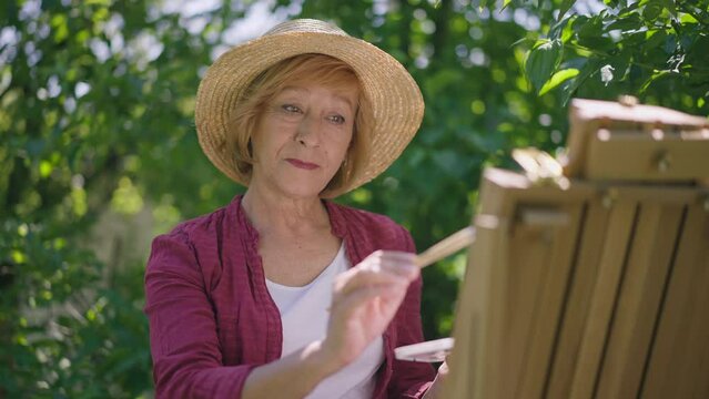 Smiling senior Caucasian woman painting on easel in slow motion standing in sunshine outdoors. Portrait of happy confident talented retiree enjoying hobby in summer spring park