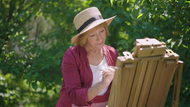 Thoughtful senior woman painting in sunshine outdoors and smiling looking at camera. Portrait of confident talented Caucasian retiree enjoying hobby posing in slow motion. Aging and lifestyle