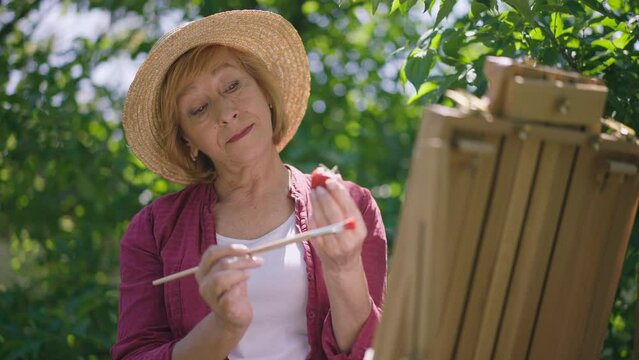 Portrait of positive talented senior woman painting strawberry in slow motion standing at easel in sunlight. Concentrated Caucasian retiree in straw hat outdoors in sunshine. Lifestyle and art concept