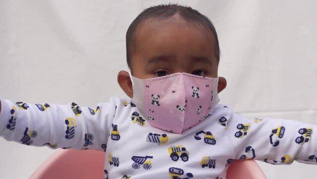 Infant baby boy is using safety mask in outdoor. The cutest toddler's baby is having fun sitting on the plastic baby chair. Portrait of a cute baby boy. Close-up view.