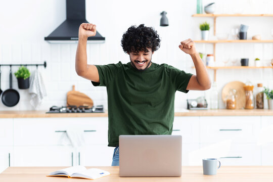 Happy young indian man looking at computer screen making yes gesture, happy with great news, received a nice message in the mail, celebrates success, smiling