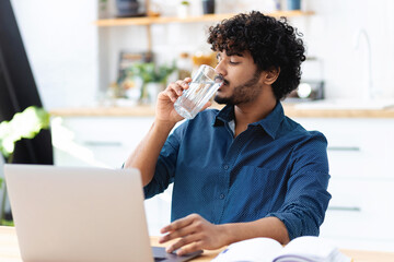 Young Indian man drinking still mineral water. Male freelancer working, using a laptop, drinks...