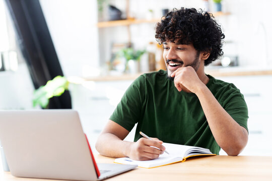 Indian Young Man Using Laptop For Watching Webinar, Studying Online And Takes Notes In A Notebook, Male Student Is Studying Online, E-learning Concept