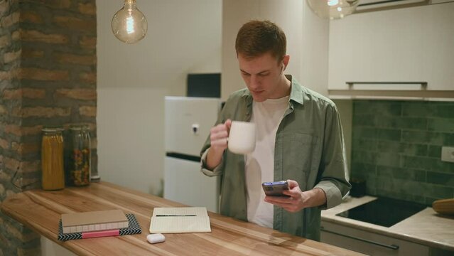 Redhead guy drinking coffee and using a smartphone and ipods  in the kitchen 