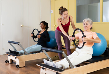 Young female pilates instructor assisting elderly woman to do exercises on reformer with flex ring and fitball. Active lifestyle concept