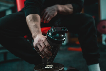 Close up on hand of unknown caucasian man holding dark supplement shaker while sitting at gym during training copy space selective focus