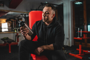Fototapeta na wymiar Front view of adult man one caucasian male athlete sitting on the bench at gym holding dark black supplement shaker while taking a brake in training bodybuilding weight lifting copy space