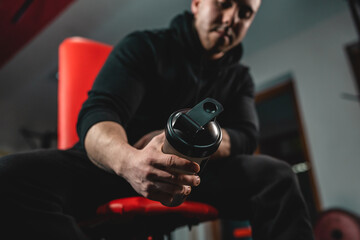 Close up on hand of unknown caucasian man holding dark supplement shaker while sitting at gym...