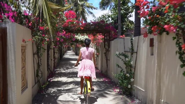 Cute girl in lovely dress riding bicycle through majestic tropical magnificent passage with pink flowers