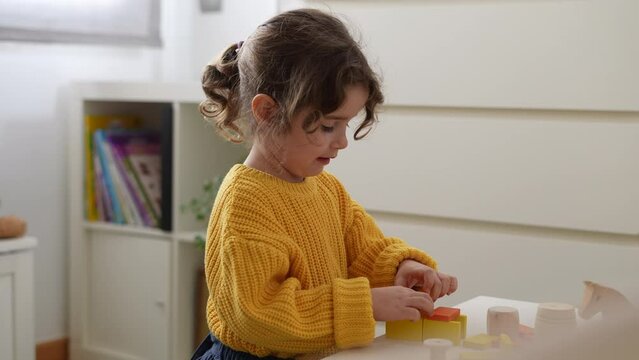 Little girl with brown hair sitting at the table in children's room playing with wooden blocks on happy weekend day