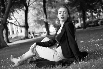 Black and white portrait of a young girl in the city park. Elegant and melancholy portrait, the girl is sitting on the grass. Beautiful woman in classic clothes