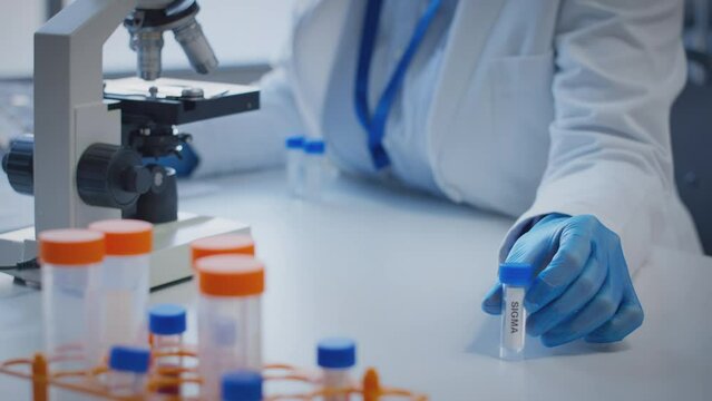 Close up of scientist wearing white coat looking through microscope and making notes whilst holding test tube which he puts on desk to show label sigma - shot in slow motion