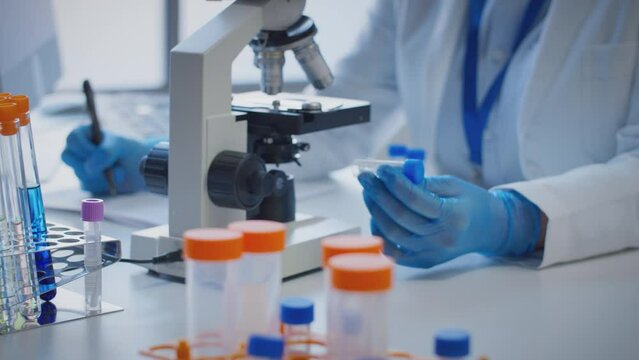 Close up of scientist wearing white coat looking through microscope and making notes whilst holding test tube which he puts on desk to show label lambda - shot in slow motion