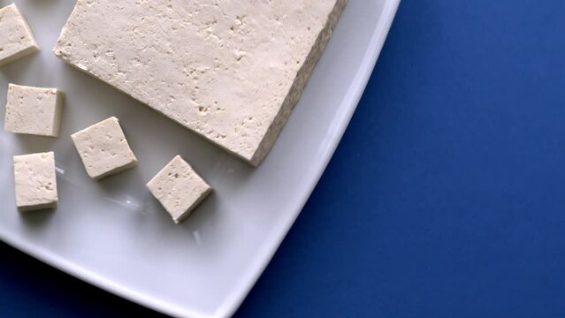 Tofu cut in cubes. Firm tofu on white plate, blue table. Vegan protein food. Table spin, top view.