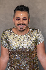 LGBT man wearing golden sequin outfit and lashes make up, posing and smiling for the camera by a gray backgrund.