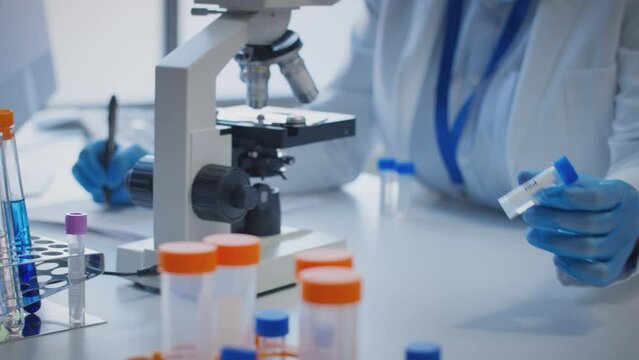 Close up of scientist wearing white coat looking through microscope and making notes whilst holding test tube which he puts on desk to show label psi - shot in slow motion