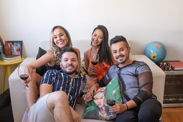 Four LGBT people drinking wine, posing and smiling at the camera, two women and two men, on the couch in a living room, during the day.