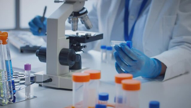 Close up of scientist wearing white coat looking through microscope and making notes whilst holding test tube which he puts on desk to show label upsilon - shot in slow motion