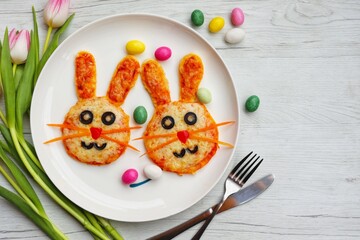 Mini Easter bunny pizzas made it from pizza crust, pizza sauce, mozzarella cheeses, black olives, carrots and red hot peppers on plate with white wood background. Art food idea for kids Easter's party