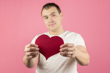 A handsome man on a pink background holds out a red paper heart in the camera, the man is out of focus. A man gives his love, a declaration of love. Valentine's Day Greeting Card