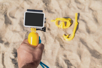 Close-up mockup Waterproof action camera with a yellow float in a male hand. On the background of the beach sand with an underwater mask with a diving snorkel. Summer beach holiday concept.