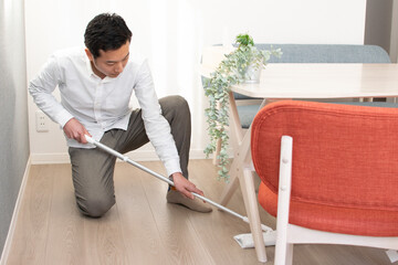 A Japenese young Asian man hoovers the floor using a vacuum cleaner in a house with a desk and two chairs