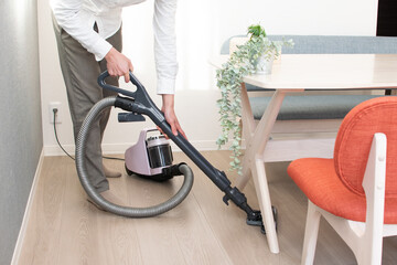 A Japenese young Asian man hoovers the floor using a vacuum cleaner in a house with a desk and two chairs
