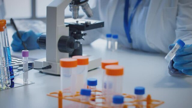 Close up of scientist wearing white coat looking through microscope and making notes whilst holding test tube which he puts on desk to show label beta - shot in slow motion