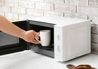 Woman putting cup into microwave oven on table near light brick wall