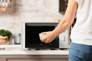Woman putting bowl with food into microwave oven in kitchen, closeup