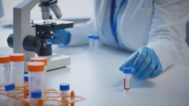 Close up of scientist wearing white coat looking through microscope and making notes whilst holding blood sample in test tube which he puts on desk to show blank label - shot in slow motion