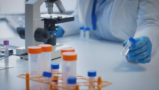 Close up of scientist wearing white coat looking through microscope and making notes whilst holding test tube which he puts on desk to show label rho - shot in slow motion